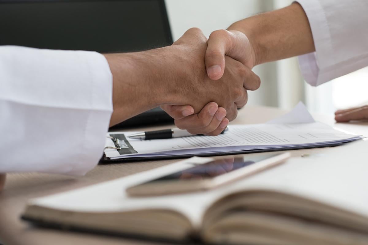 Two people shaking hands over a desk with a clipboard, phone, and open book on it.